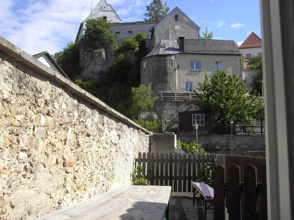 Blick über die Terrasse mit Tisch und Bank auf die alte Stadtmauer und das Schloss auf dem Felsen darüber.