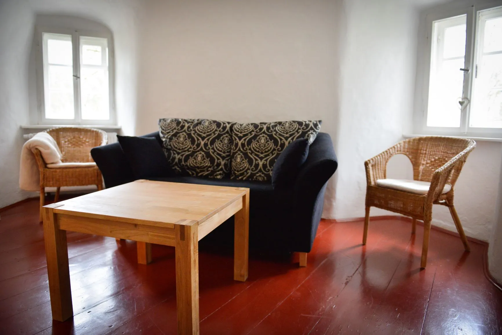 Der Wohnraum im Untergeschoss: Sofa, zwei Korbsessel und ein Holztisch auf rotem Dielenboden.